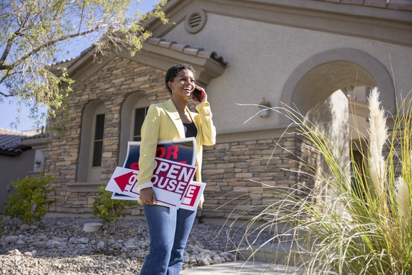 Person wearing a yellow jacket, standing outside of a house on a cell phone, holding a open house and for sale sign.