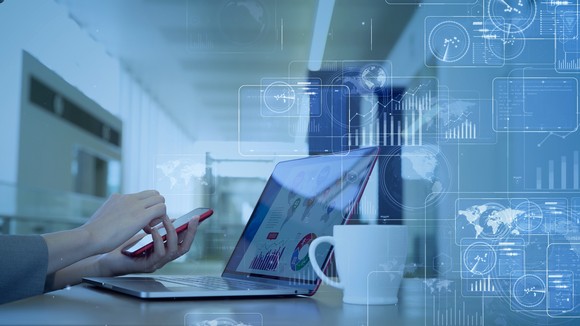 Hands operate a phone next to a laptop or cup of coffee. 