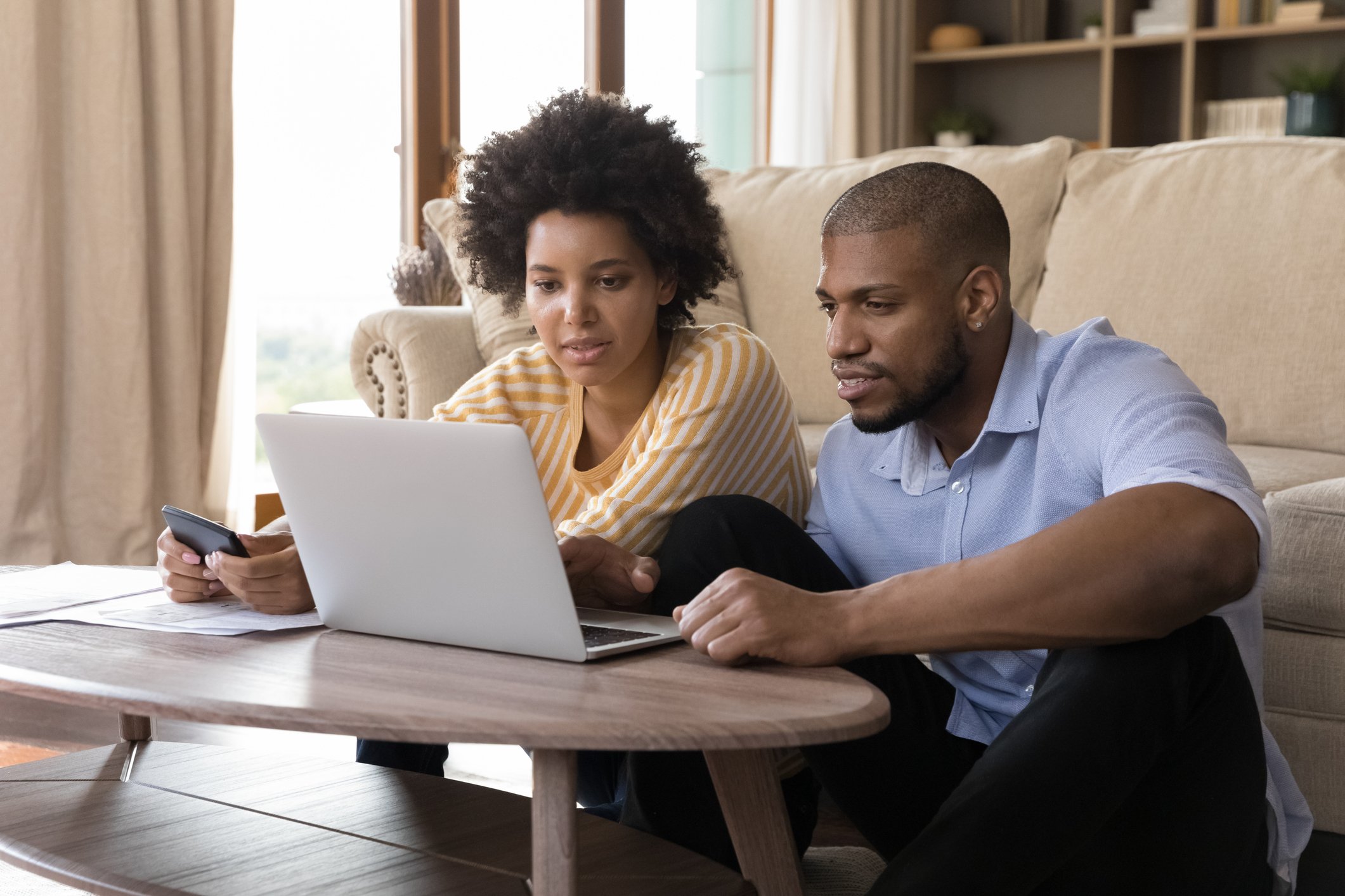Two people sit on the floor at a coffee table and study something on a laptop.