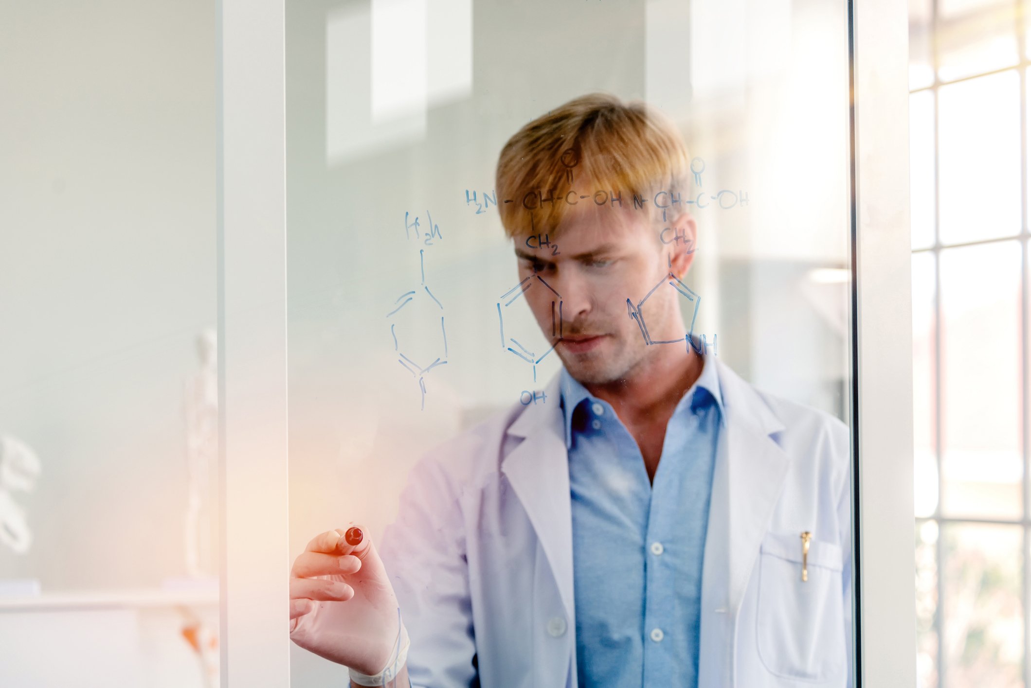 A scientist draws schematics of molecules on a window with a dry-erase marker while standing in a conference room looking perplexed.