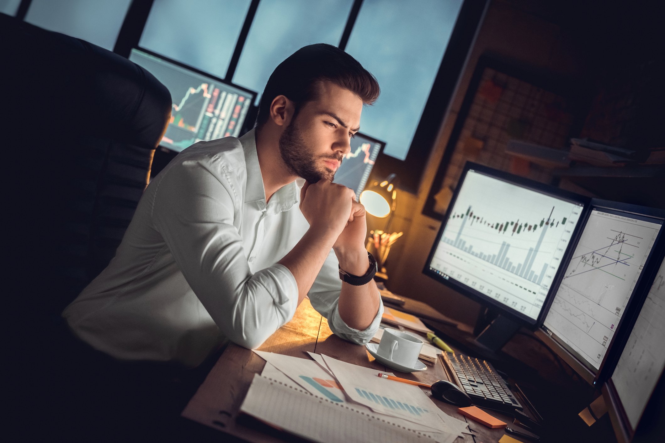 Tech investor looking concerned at his desk before computer monitors. 