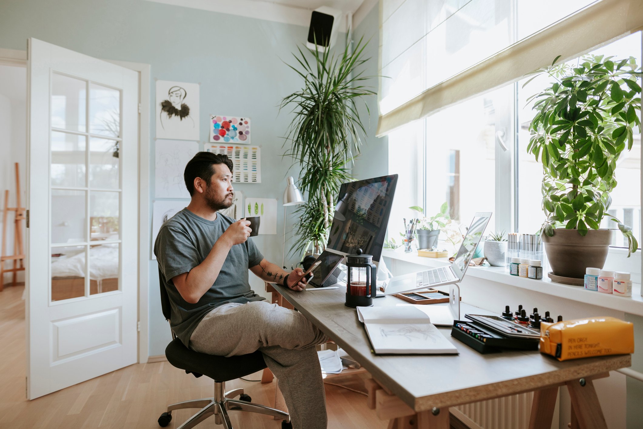 A person holding a drink and working in an open, airy, home office with plants.