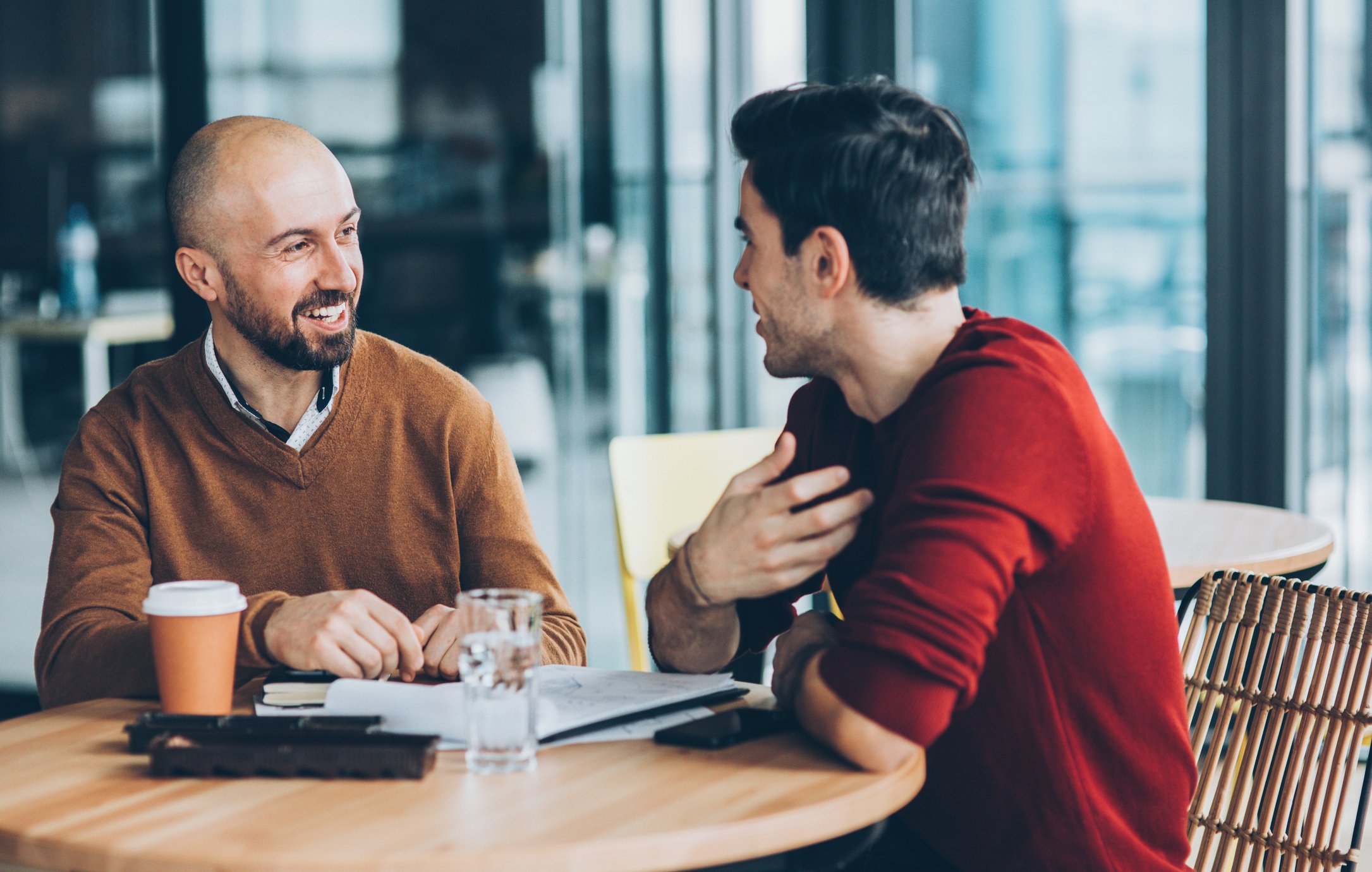 Two gentlemen are smiling while sitting and talking at a table outside of a cafe. 