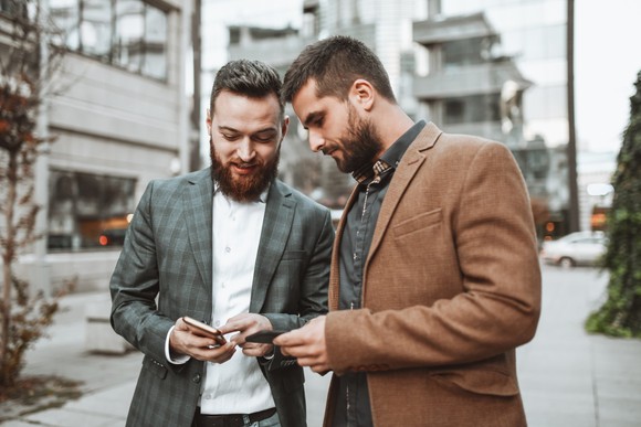 Two investors look at something on their phones on a city sidewalk.