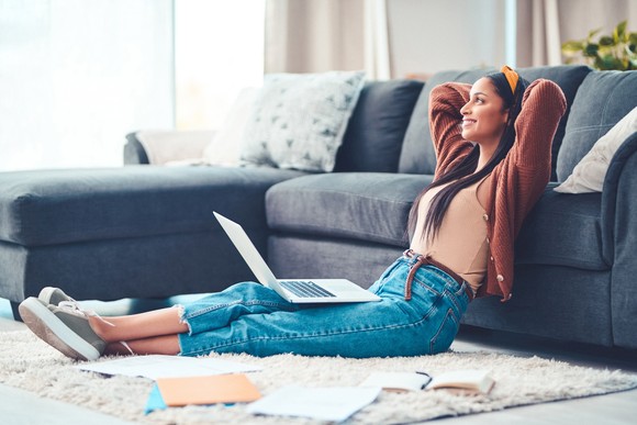 A young adult sits on the floor and leans against the couch smiling with a laptop on her lap.