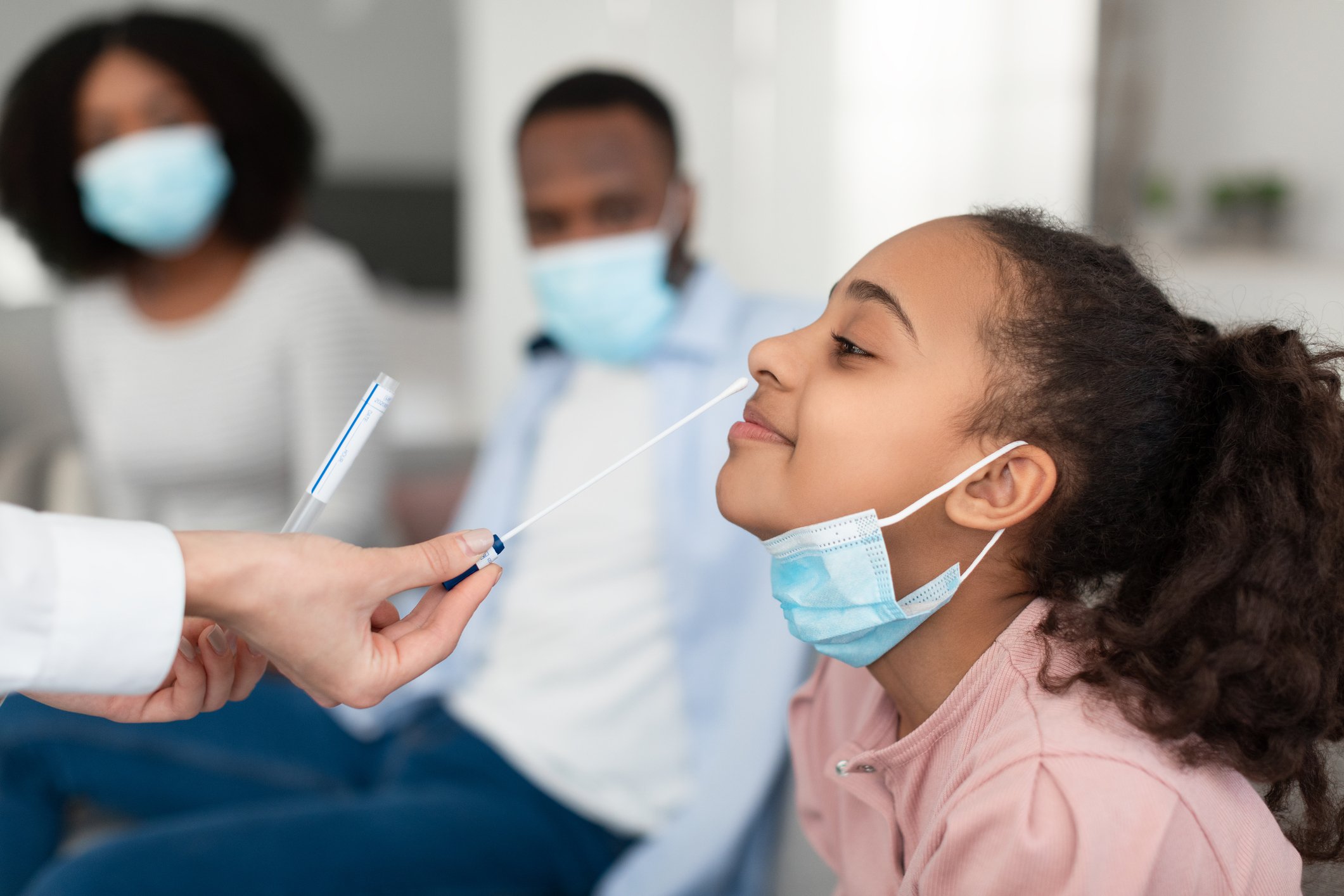 A child having their nose swabbed for a COVID test while parents watch in the background.
