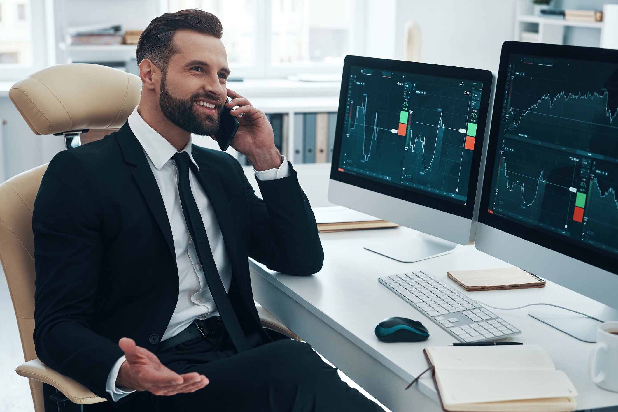 Man in a suit talking on the phone, while price charts are displayed on two computers on his desk.