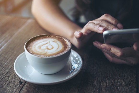 A person's hands type something on a phone near a cup of coffee on a table.
