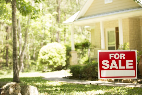 A home on a spring day with a for sale sign in green grass.