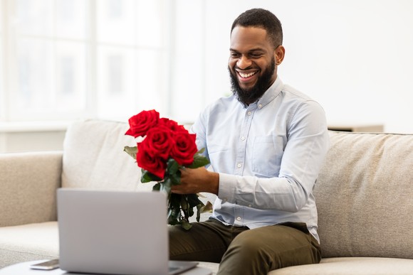 An investor sits on a couch holding a bouquet of red roses and smiles into a laptop.