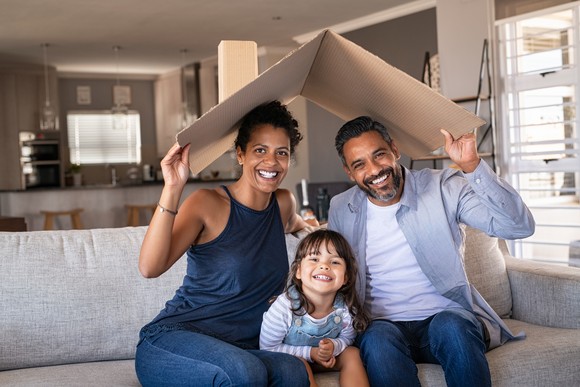 Three people on a couch holding a cardboard flap that looks like a roof over their heads.