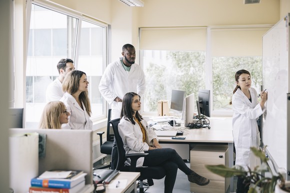 A scientist writes on a whiteboard as coworkers seated in an office look on.