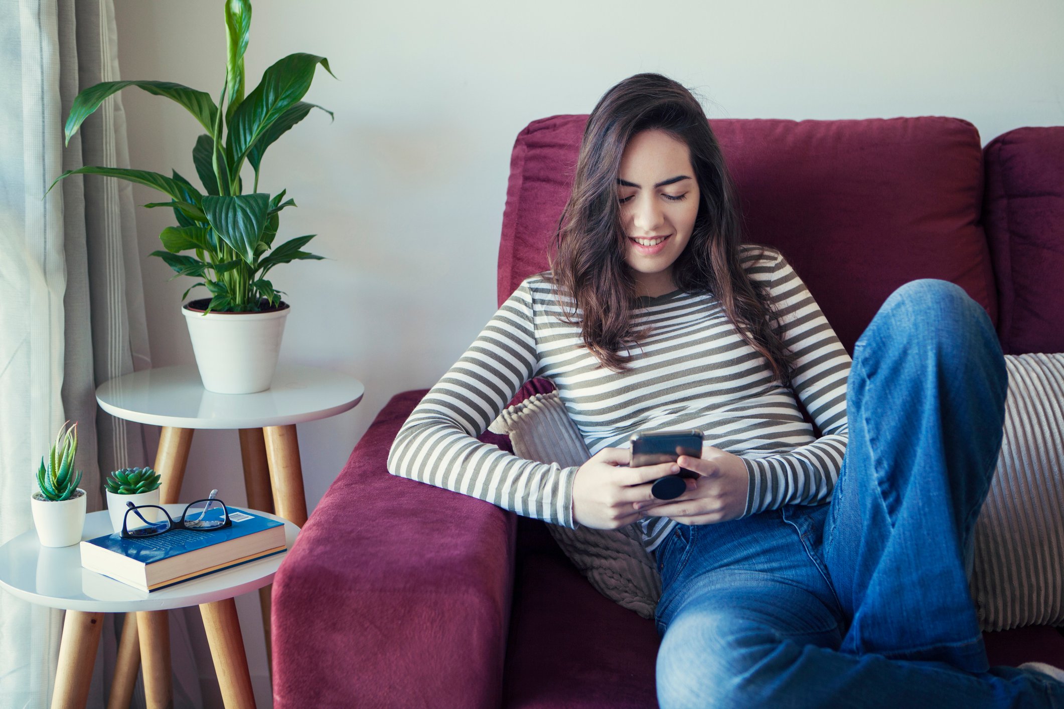 A young adult sits on a couch smiling and using a smartphone.