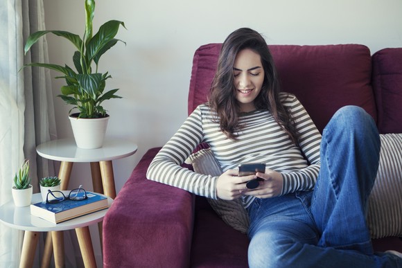 A young adult sits on a couch smiling and using a smartphone.