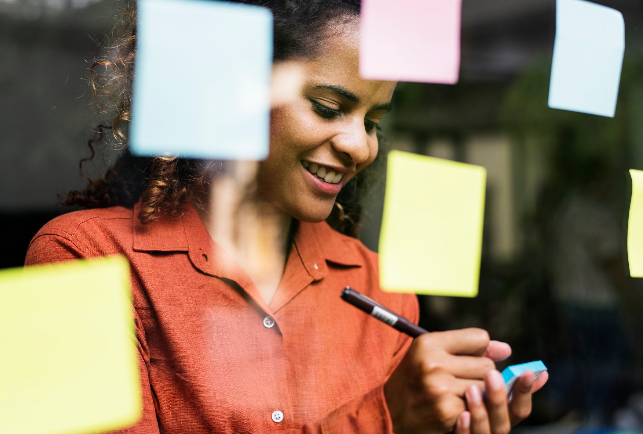 Person writing on a sticky note in front of a clear wall with several sticky notes attached to it. 