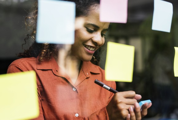 Person writing on a sticky note in front of a clear wall with several sticky notes attached to it. 