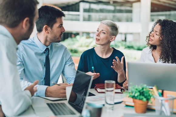 Four businesspeople sit around a table talking with notepads and an open laptop.