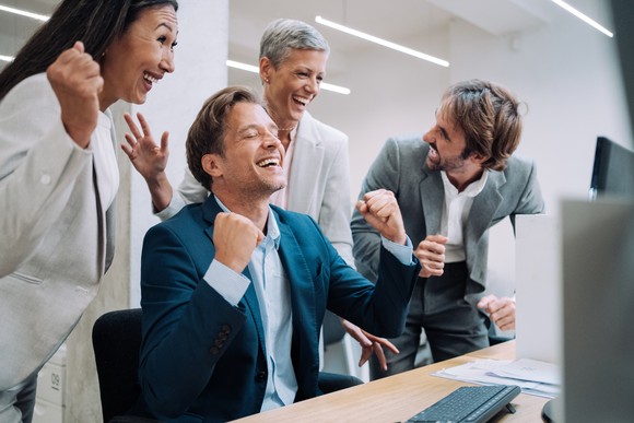Happy business people celebrating with each other around a computer screen at a desk.