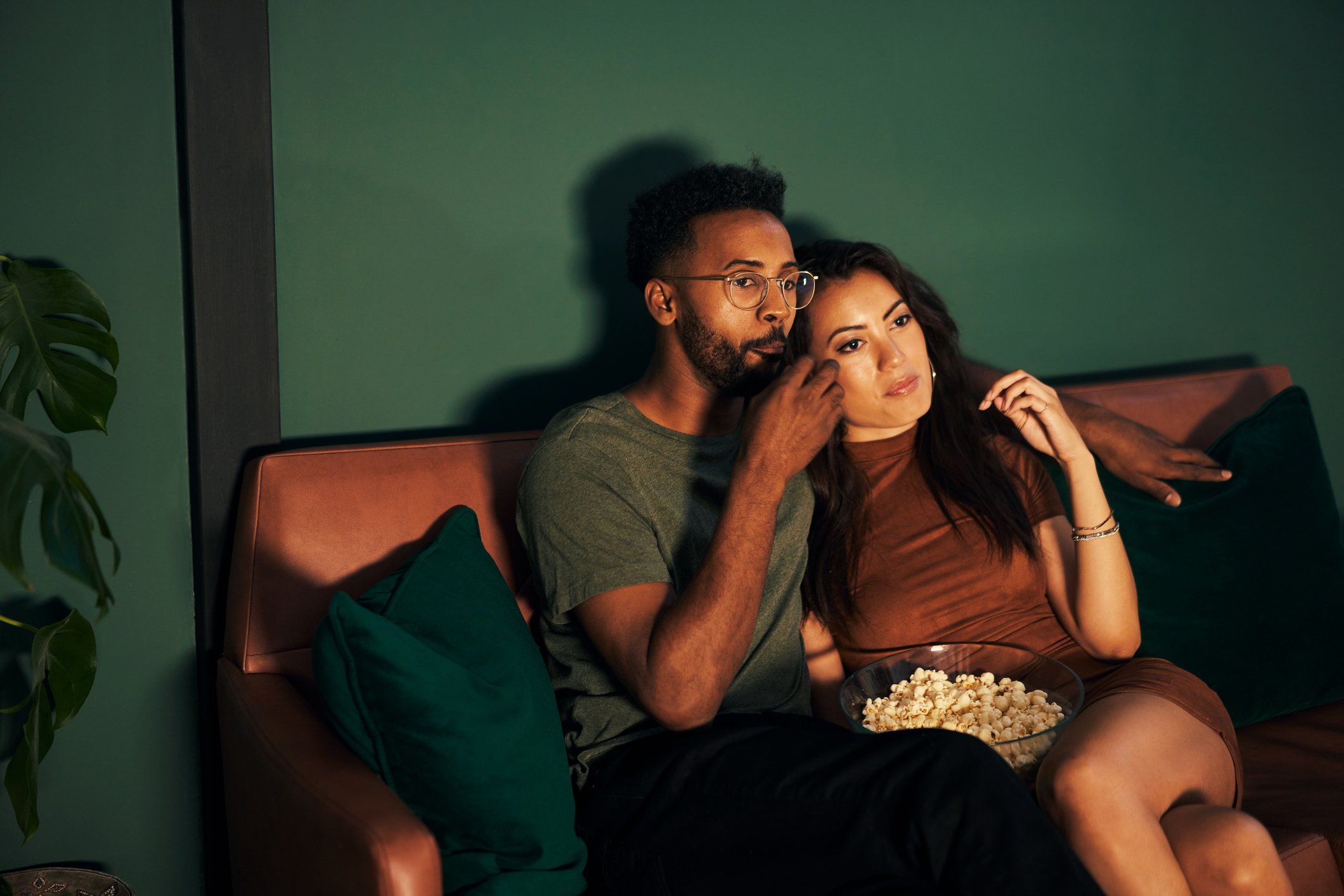 Young couple seated on a couch, eating popcorn and watching TV.