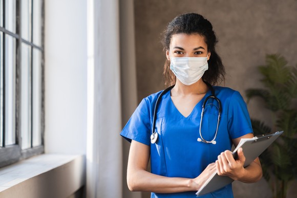 A healthcare professional in uniform and mask, holding a clipboard.