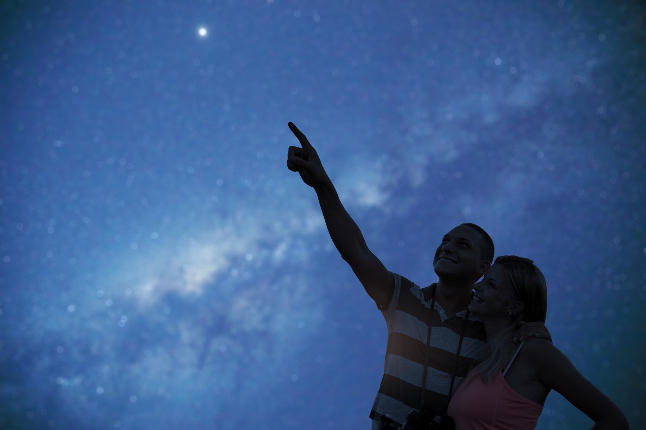 Two people stand close together outdoors and one of them points to a star in the night sky.