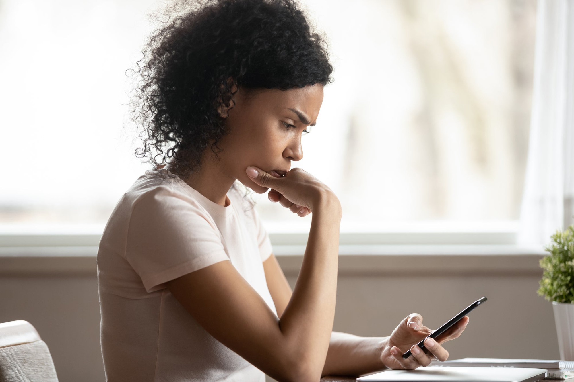 A woman sits at a table and looks at her phone with a concerned look on her face.