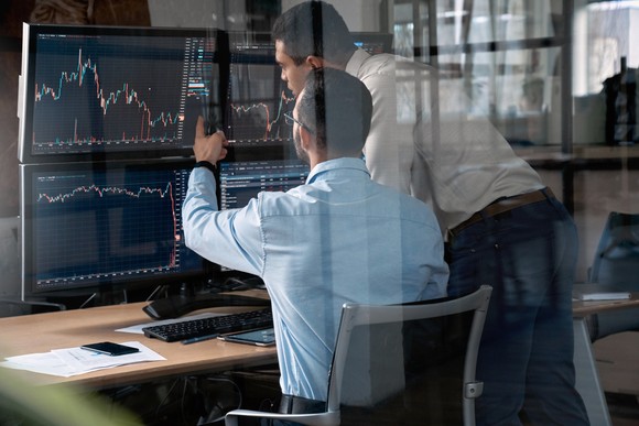 Two traders looking at charts on multiple screens.