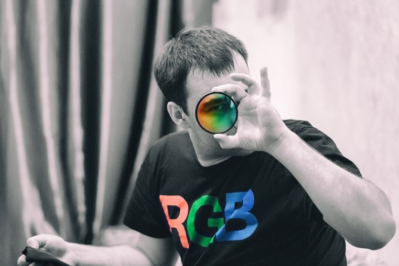 A person wearing a shirt with the letters RGB holds up a lens that brings color to an otherwise greyscale image.