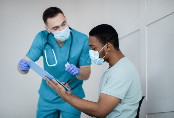 A healthcare worker holds out forms to be signed by a patient in a medical setting prior to vaccination.