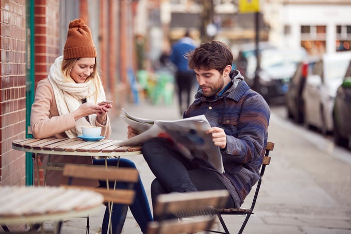 Two people at a table reading a newspaper and phone.