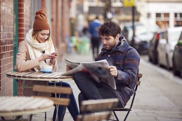 Two people at a table reading a newspaper and phone.