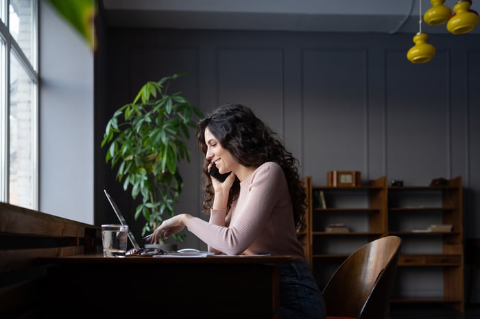 A smiling person at a desk in a home office talks into a smartphone while scrolling on a laptop.