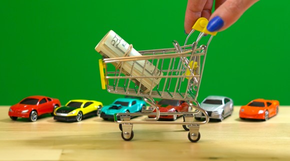 Miniature shopping cart filled with money, next to miniature cars.