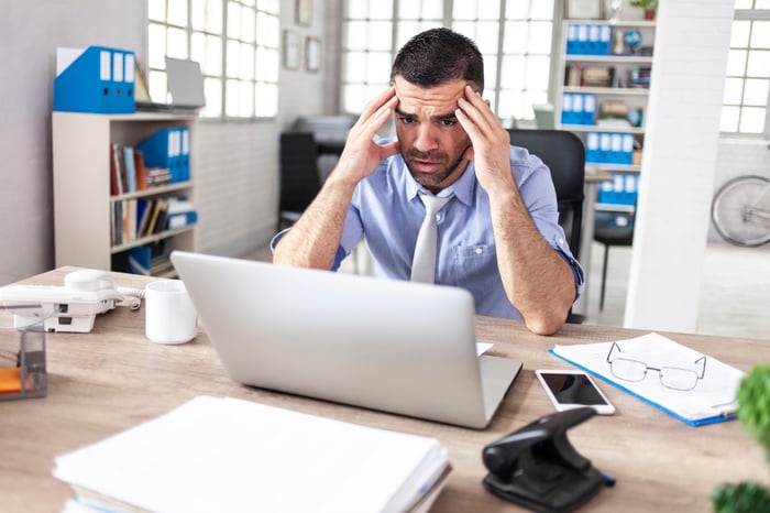 A person in an office holding their head in frustration while looking at a laptop.