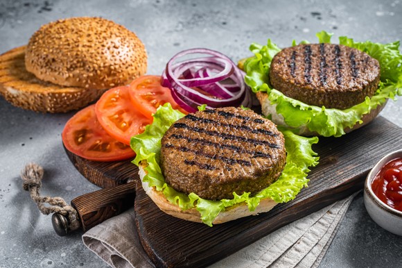 Plant-based meatless burgers with vegan grilled patties, tomato, and onion on a wooden serving board.