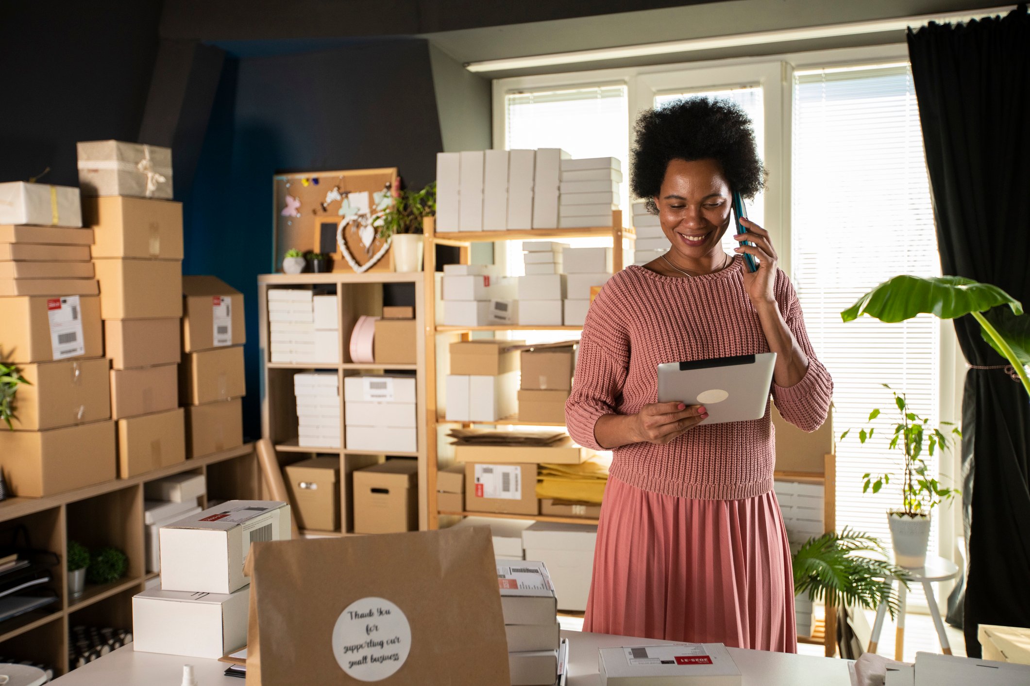 Young woman on phone looks at tablet in store room with cardboard boxes. 
