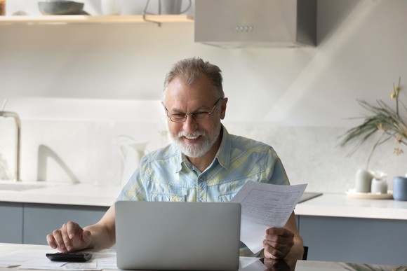 Middle aged person sitting at desk  holding papers while smiling and looking at computer