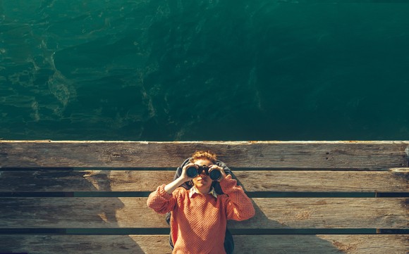 Person lying down on a dock alongside water and looking through binoculars.