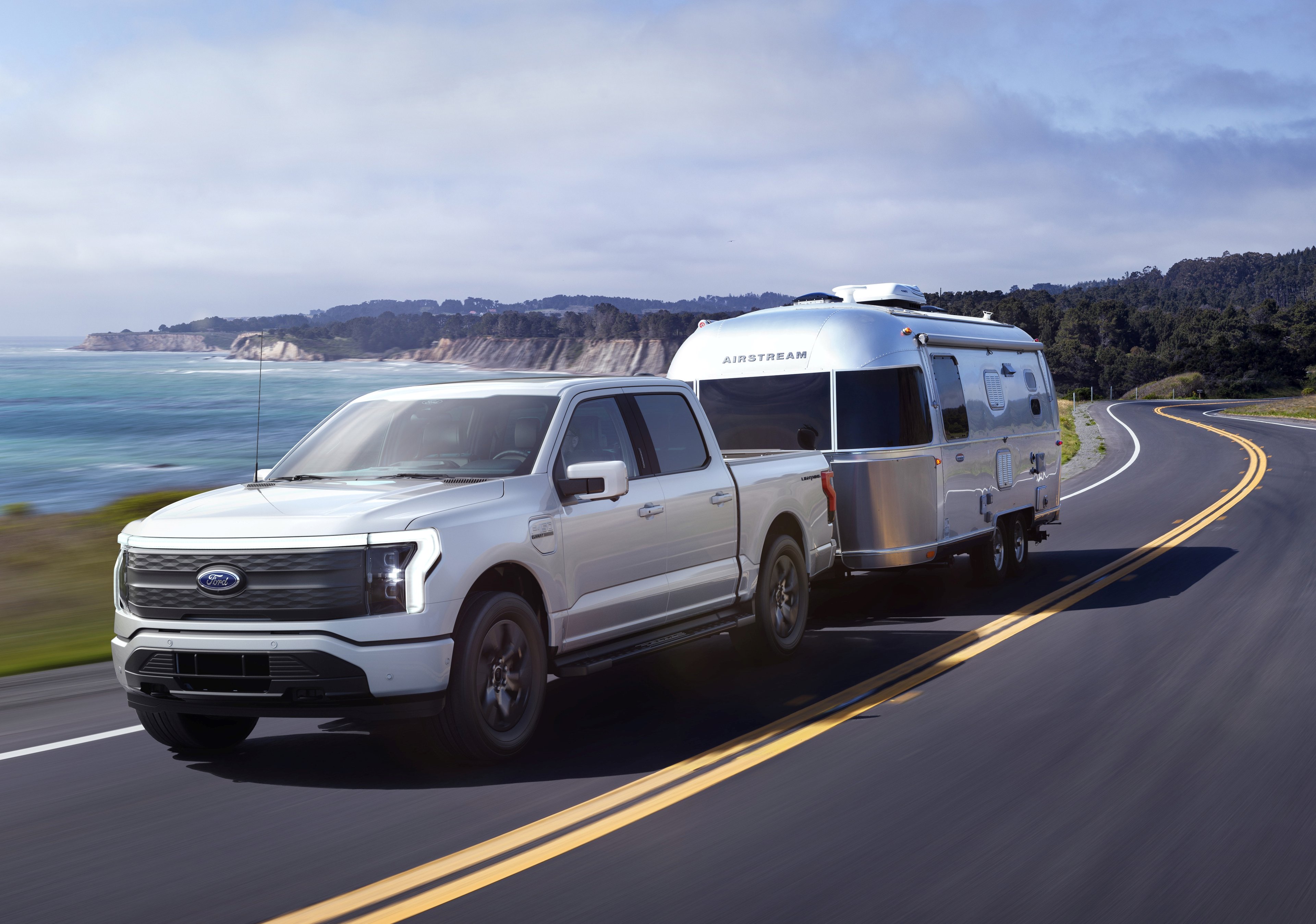 Ford F-150 Lightning towing a trailer on a coastal highway with the ocean in the background.
