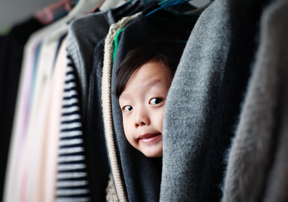 A child peeks out while hiding between hanging clothes in a closet.