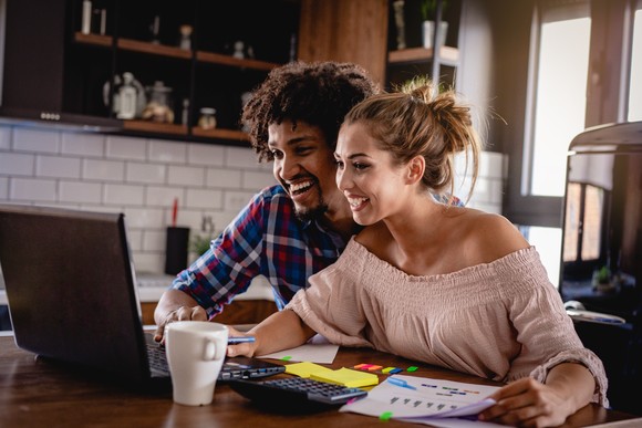 Two investors smile brightly as they look at a laptop in a living room setting. 