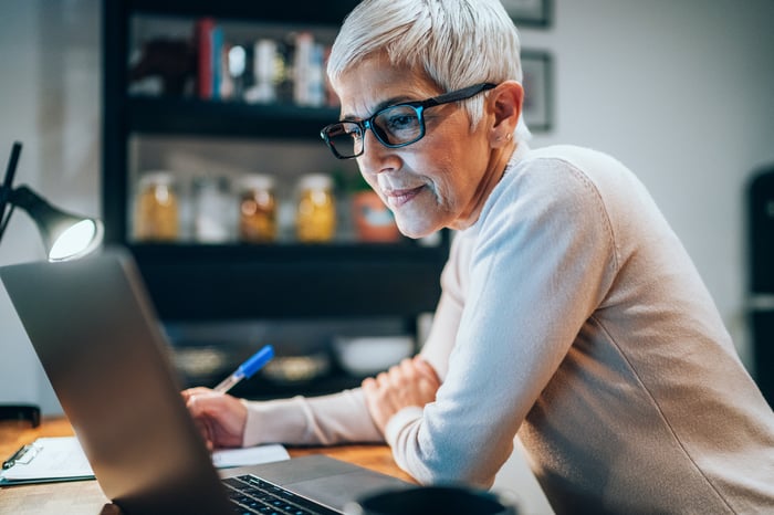 Serious senior looking at laptop and writing note.