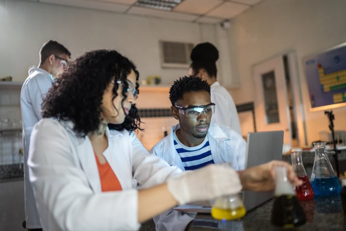 Four scientists work in a laboratory together as one holds a pair of beakers at a laboratory bench.