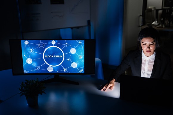 Person gazing at a laptop, next to a video display with a "Blockchain" graphic.