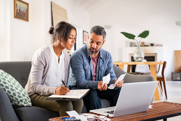 Two people sitting on a couch looking at documents and a laptop.