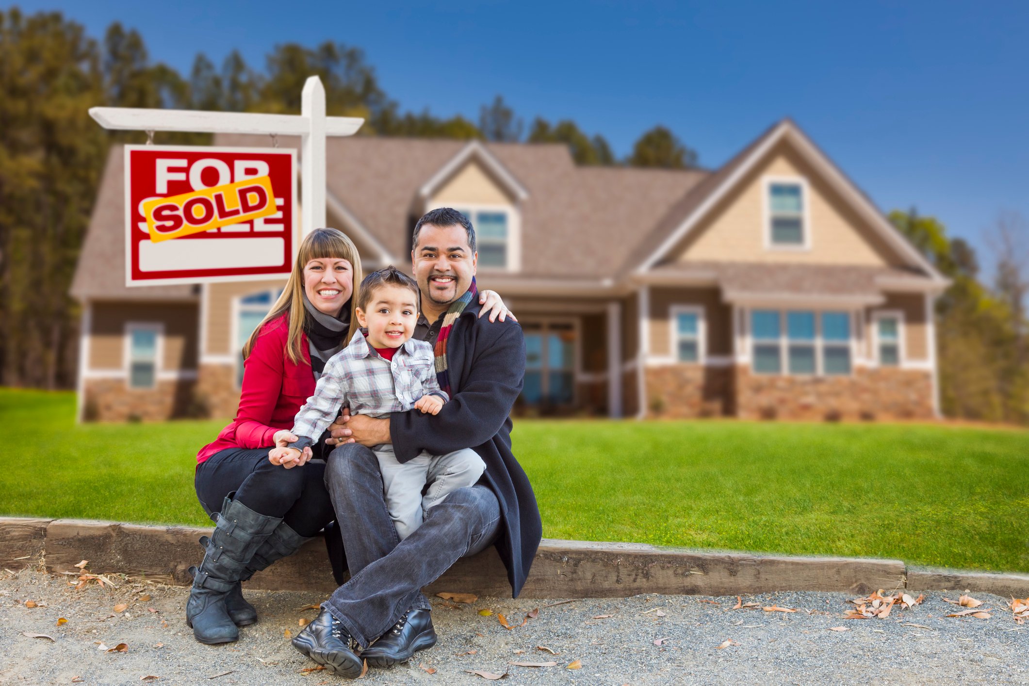 Two adults and one child, all smiling, in front of a real estate sign in a home's front yard.