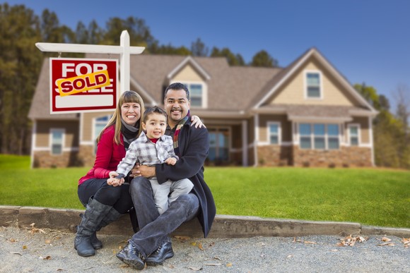 Two adults and one child, all smiling, in front of a real estate sign in a home's front yard.