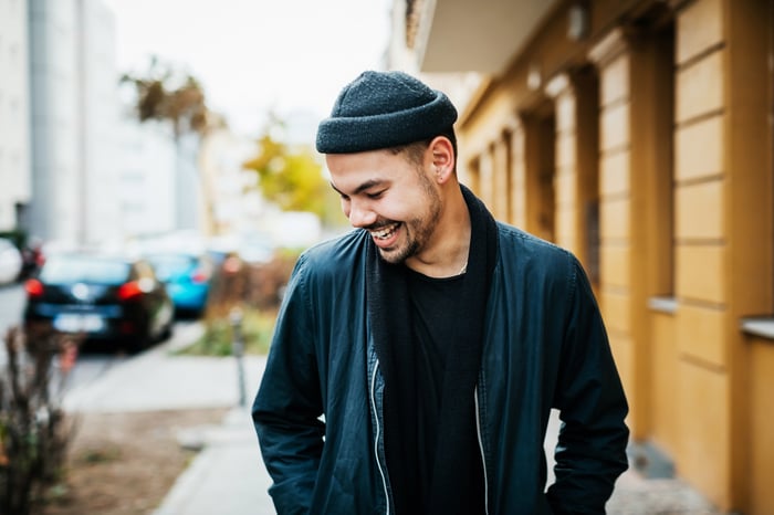 Young adult wearing beanie laughs while walking on urban sidewalk.
