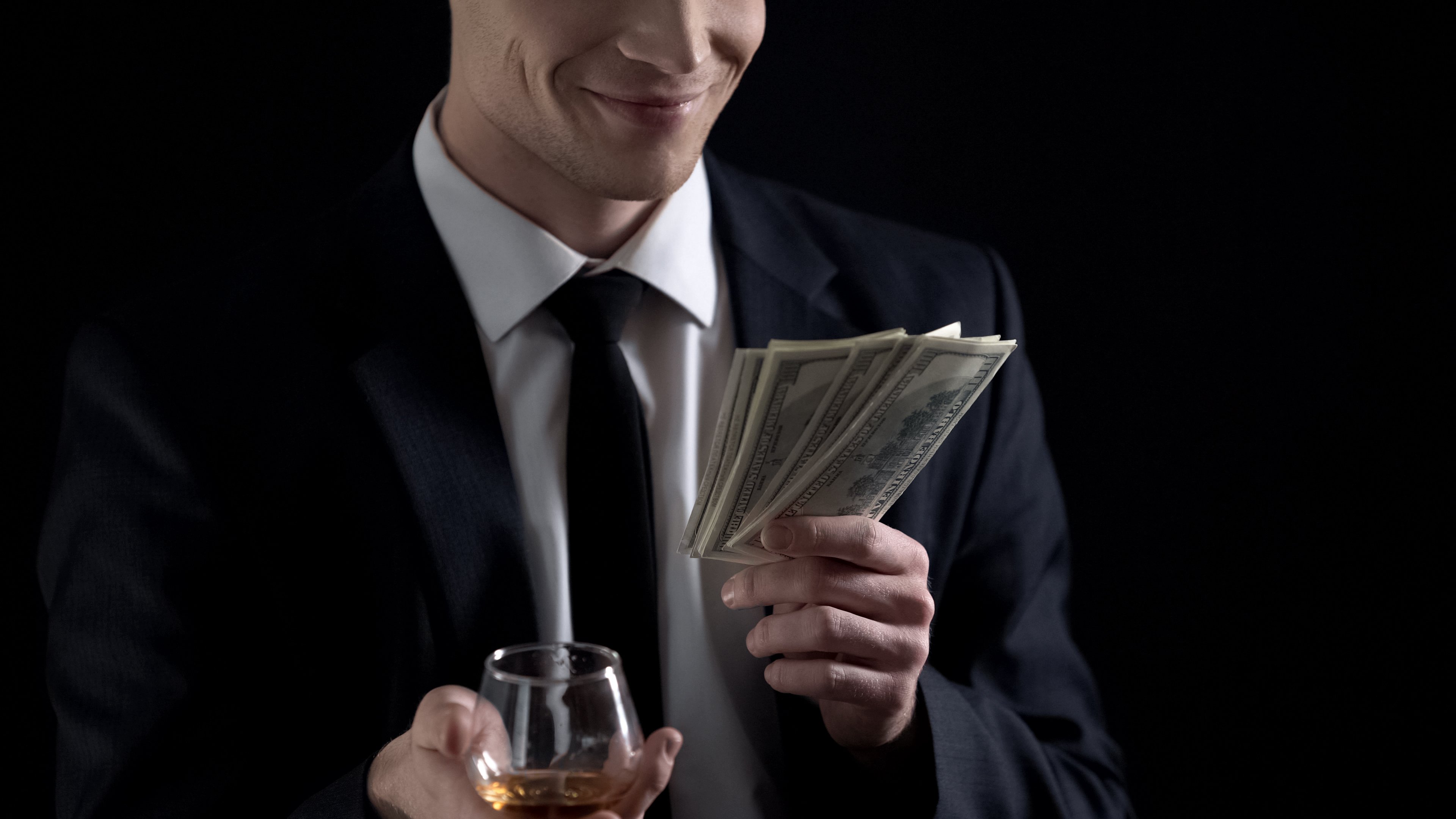 Investor in a suit holds cash in one hand a brandy glass in the other. 
