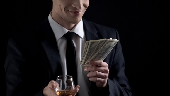 Investor in a suit holds cash in one hand a brandy glass in the other. 